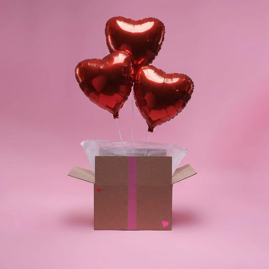 Box cadeau ouverte avec trois ballons cœur rouges flottants, mise en scène sur fond rose.