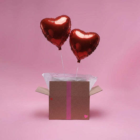 Duo de ballons cœur rouges flottant au-dessus d’une boîte cadeau décorée, fond rose pastel