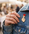 Pin's émaillé "Finger Heart" (cœur avec les doigts) de haute qualité, rouge et beige, épinglé sur le col d'une veste en jean gris délavé.