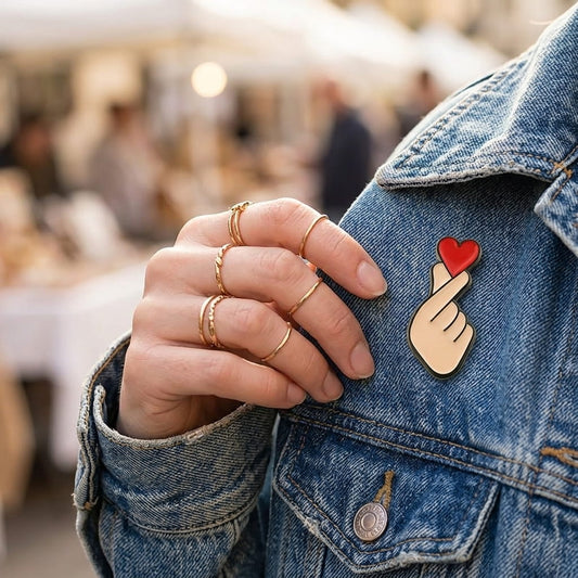 Pin's émaillé "Finger Heart" (cœur avec les doigts) de haute qualité, rouge et beige, épinglé sur le col d'une veste en jean gris délavé.