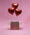 Ensemble de trois ballons cœur rouges en aluminium dans une box décorative sur fond rose.
