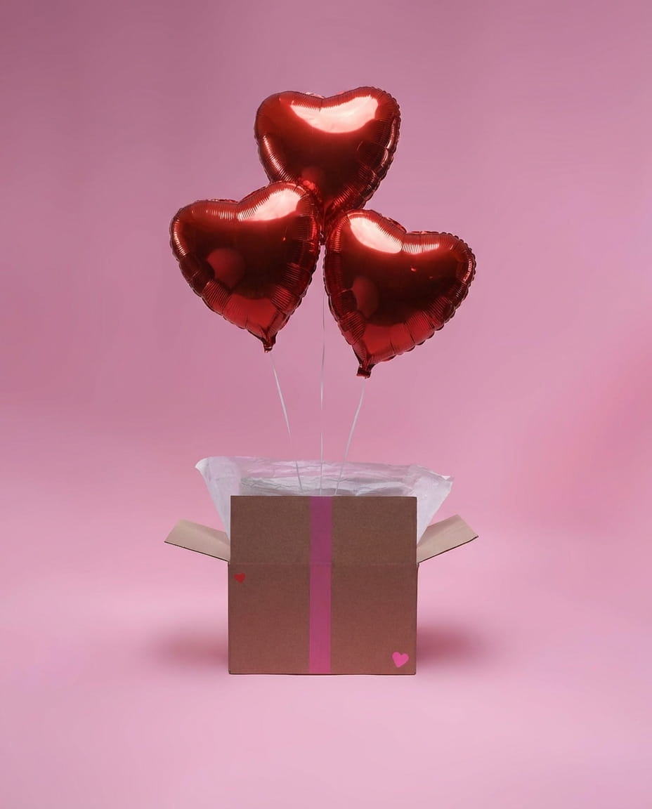 Ensemble de trois ballons cœur rouges en aluminium dans une box décorative sur fond rose.