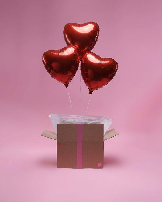 Ensemble de trois ballons cœur rouges en aluminium dans une box décorative sur fond rose.