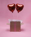 Deux ballons cœur rouges gonflés à l’hélium sortant d’une box cadeau sur fond rose
