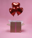 Trois ballons cœur rouges gonflés à l’hélium sortant d’une box cadeau sur fond rose.