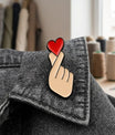Main de femme portant des bagues fines en argent, montrant le pin's Finger Heart fixé sur sa veste en jean bleu clair. Arrière-plan flou avec plantes vertes et guirlande lumineuse.