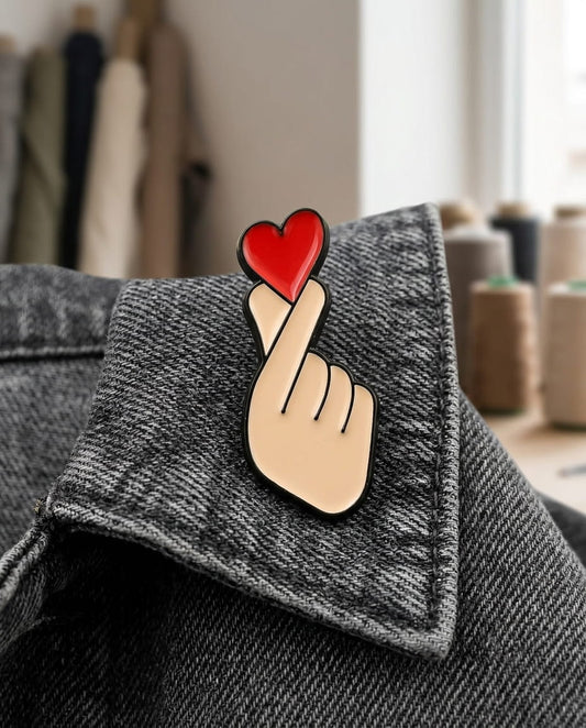 Main de femme portant des bagues fines en argent, montrant le pin's Finger Heart fixé sur sa veste en jean bleu clair. Arrière-plan flou avec plantes vertes et guirlande lumineuse.