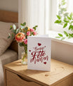 Carte de vœux blanche avec calligraphie rouge "Bonne Fête Maman" et petits cœurs, enveloppe rouge en arrière-plan.