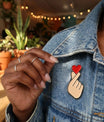 Plan détaillé sur une main de femme tenant le col de sa veste en jean bleu pour mettre en valeur le pin's Finger Heart (cœur avec les doigts). Bijoux fins et bagues en argent visibles.