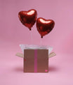 Duo de ballons cœur rouges flottant au-dessus d’une boîte cadeau décorée, fond rose pastel