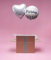 Duo de ballons argent dont un cœur et un ballon Happy Birthday sortant d’une box décorative fond rose