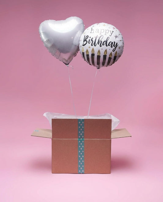 Duo de ballons argent dont un cœur et un ballon Happy Birthday sortant d’une box décorative fond rose