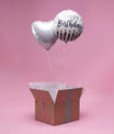 Box cadeau ouverte avec papier de soie laissant s’élever deux ballons argentés cœur et Happy Birthday sur fond rose