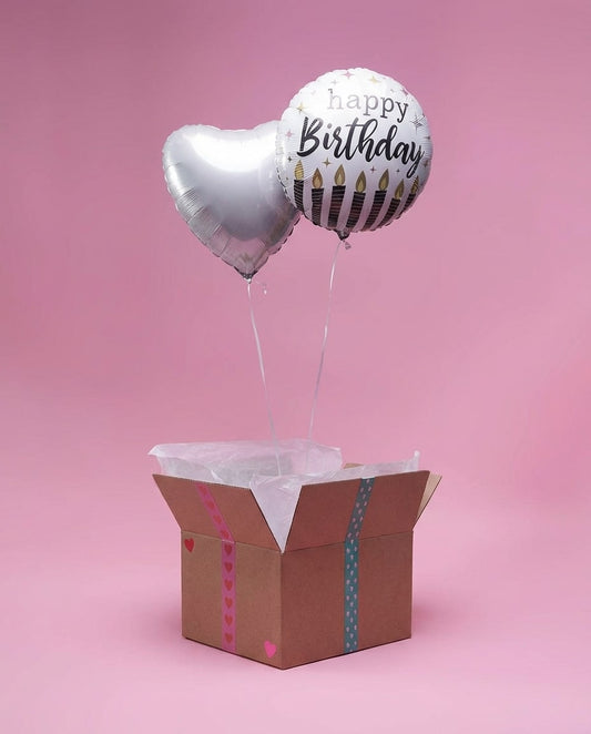 Box cadeau ouverte avec papier de soie laissant s’élever deux ballons argentés cœur et Happy Birthday sur fond rose