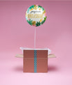 Ballon Tropical Joyeux Anniversaire flottant au-dessus d’une boîte cadeau rose