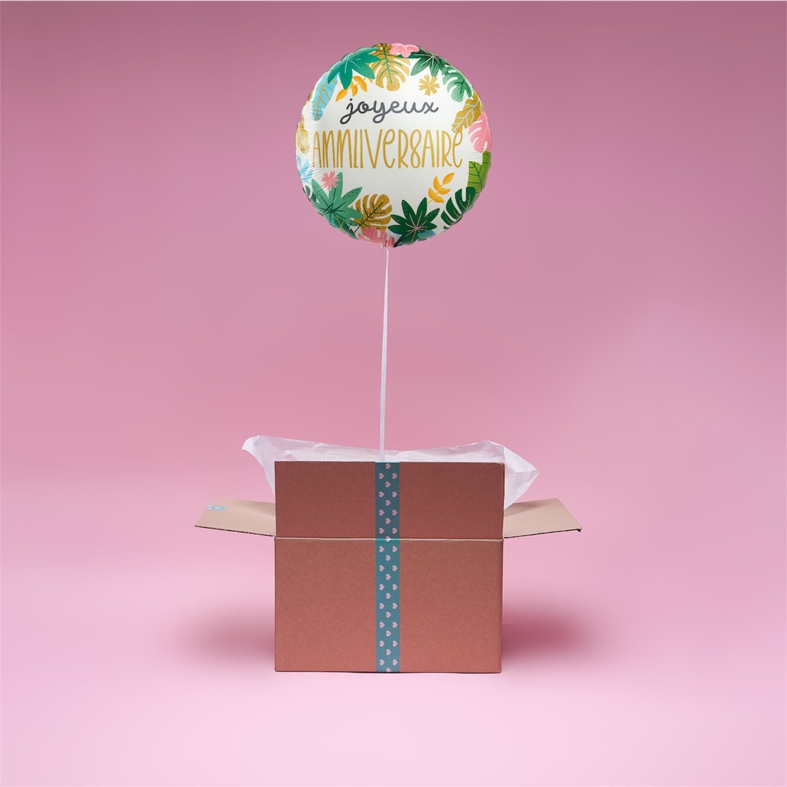 Ballon Tropical Joyeux Anniversaire flottant au-dessus d’une boîte cadeau rose