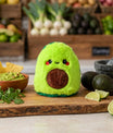 Bouillotte peluche avocat posée sur une planche en cuisine avec avocats et guacamole