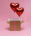 Duo de ballons cœur rouges flottant au-dessus d’une boîte cadeau décorée, fond rose pastel