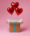 Trois ballons cœur rouges gonflés à l’hélium sortant d’une box cadeau sur fond rose.