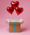 Box cadeau ouverte avec trois ballons cœur rouges flottants, mise en scène sur fond rose.