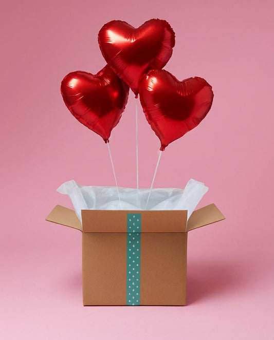Box cadeau ouverte avec trois ballons cœur rouges flottants, mise en scène sur fond rose.