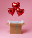 Ensemble de trois ballons cœur rouges en aluminium dans une box décorative sur fond rose.