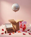 Coffret cadeau festif avec ballon hélium, oursons chocolatés et sachets de bonbons en forme de cœurs sur un fond rose poudré.